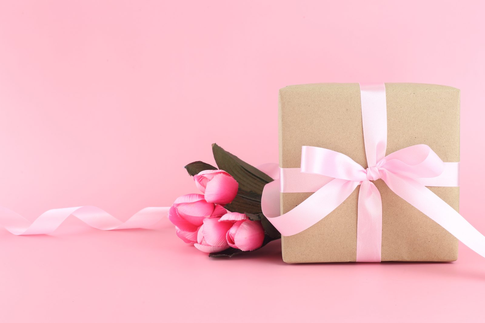 brown paper gift box with pink satin curly ribbon bow on pastel pink background. flat lay mother day, father day, valentines day, birthday concepts with copy space.